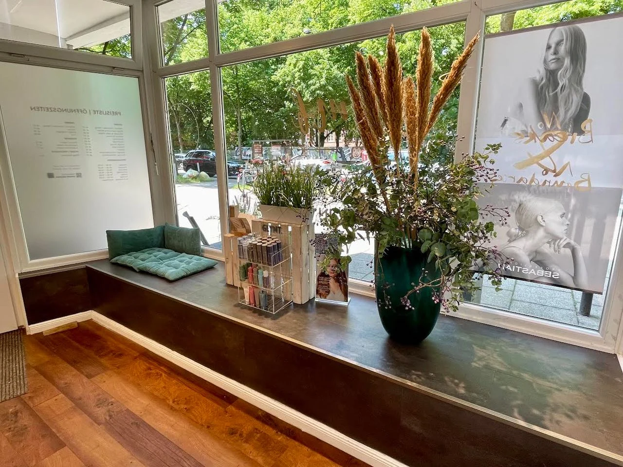 Long bench by the shop window with petrol-coloured cushions and pampas grass, a tree-lined street outside